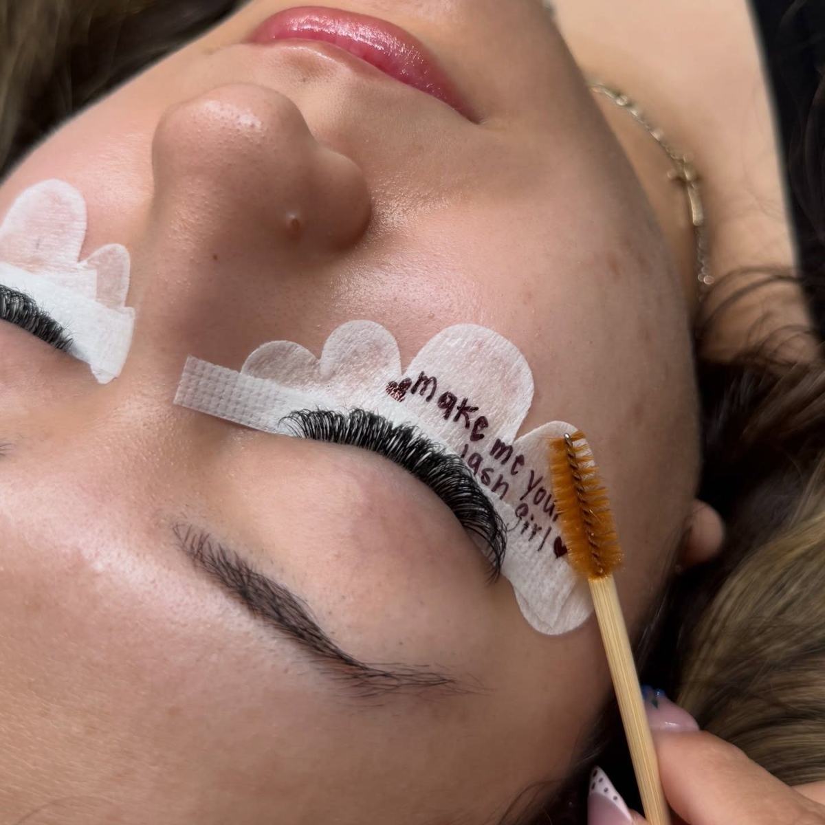 Under-eye tape with 'make me your lash girl' written on it in marker, lashes being brushed through